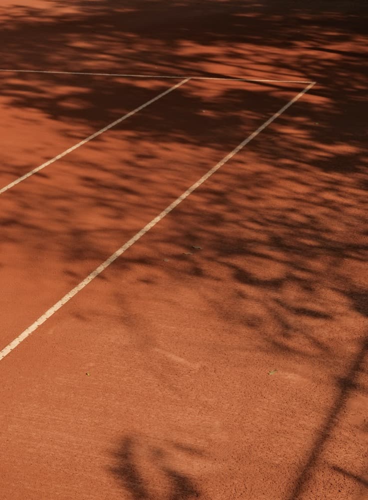 Crushed-brick clay surface of Court B marked with crisp white lines under dappled shadow